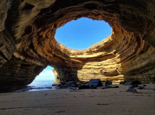 A sandy beach with a large rock formation featuring a circular opening, revealing a clear blue sky above.