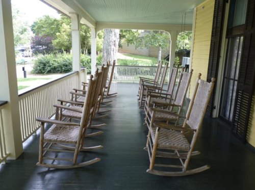 A row of wooden rocking chairs on a porch, surrounded by greenery and sunlight.