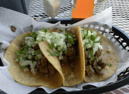 Three tacos filled with seasoned meat, topped with chopped onions and cilantro, served in a basket.