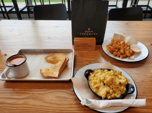 A table with a cup of tomato soup, grilled cheese, and a skillet of mac and cheese, alongside chicken wings and a sign.