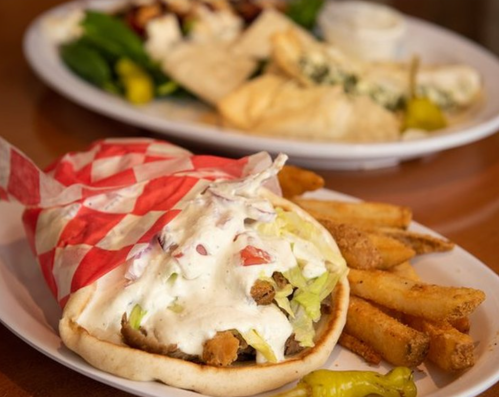 A pita filled with chicken, lettuce, and sauce, served with fries on a plate, with another dish in the background.