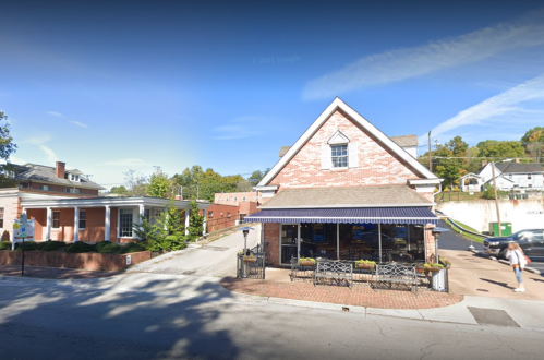 A brick building with a blue awning, surrounded by trees and a sidewalk, on a sunny day.