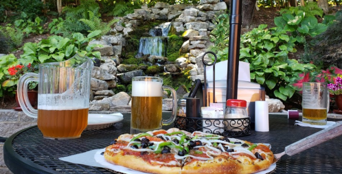 A table set outdoors with pizza, two beers, and a scenic waterfall in the background surrounded by greenery.