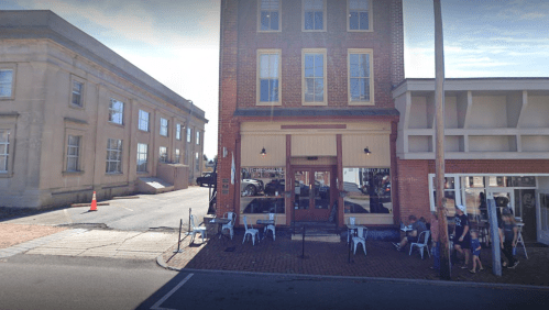A brick building with outdoor seating, located on a street corner, with people sitting outside and a nearby parking area.