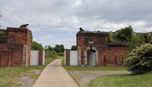 A pathway leads through two brick gate structures, with greenery on either side and a cloudy sky above.