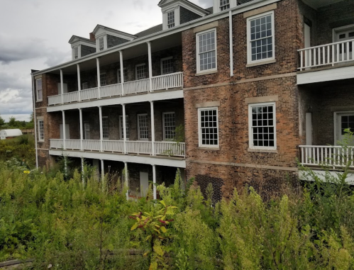 Abandoned brick building with multiple balconies, surrounded by overgrown vegetation and cloudy skies.