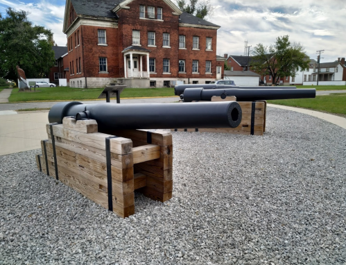 Two historic cannons mounted on wooden platforms, set against a backdrop of a brick building and cloudy sky.