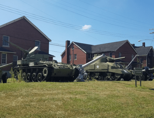 Two military tanks displayed on grass near brick buildings, with clear blue skies above.