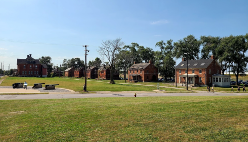 Historic brick buildings line a grassy area under a clear blue sky, with trees in the background and a few people walking.