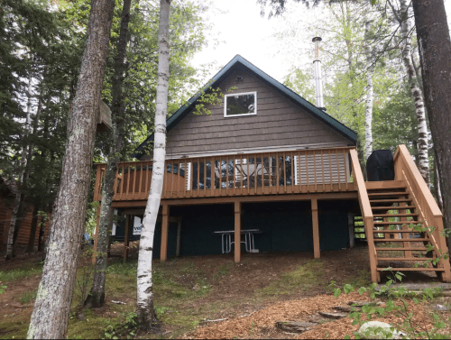 A cozy cabin with a wooden deck, surrounded by trees and greenery, featuring a staircase leading to the entrance.