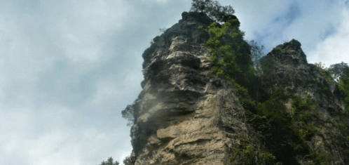 A towering rock formation surrounded by greenery under a cloudy sky.