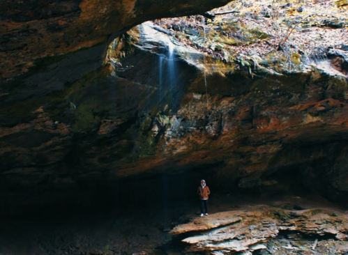 A person stands beneath a rocky overhang with a small waterfall cascading down in a serene, natural setting.