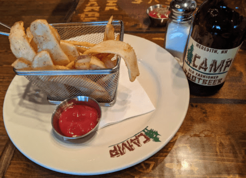 A plate of fries in a metal basket with ketchup, alongside a bottle of root beer on a wooden table.
