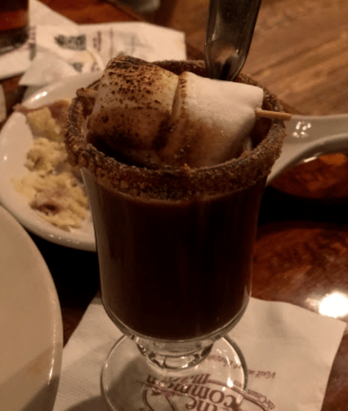 A glass of hot chocolate topped with a toasted marshmallow and a graham cracker rim, served on a wooden table.