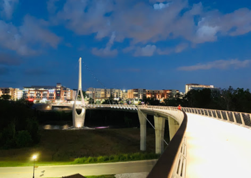 A modern bridge illuminated at night, with a city skyline and clouds in the background.