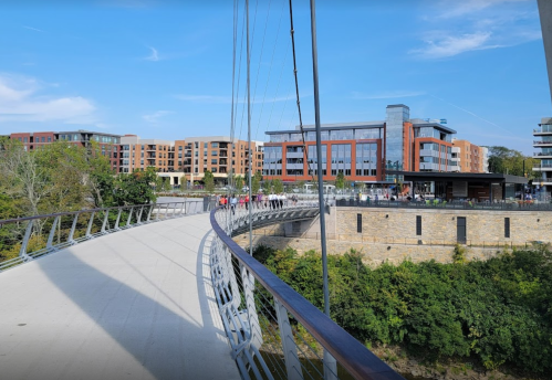 A curved pedestrian bridge leads to modern buildings, surrounded by greenery and a clear blue sky.