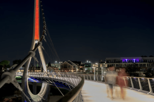 A illuminated bridge at night with blurred figures walking, surrounded by buildings and lights in the background.