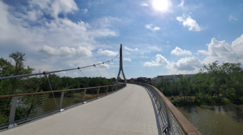 A modern bridge spans a river, surrounded by greenery and under a bright blue sky with fluffy clouds.