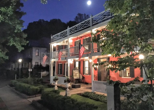 A charming red house with balconies, adorned with American flags, under a moonlit sky. Lush greenery surrounds the scene.