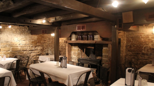 Cozy restaurant interior with stone walls, wooden beams, tables set with white linens, and a fireplace in the background.