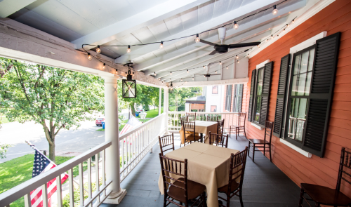 A charming porch with tables, chairs, and string lights, overlooking a tree-lined street and an American flag.