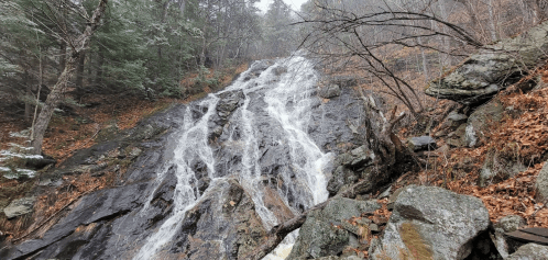 A cascading waterfall flows down a rocky slope, surrounded by trees and autumn leaves. Mist adds a serene atmosphere.