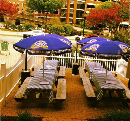 Outdoor dining area with wooden picnic tables under blue umbrellas, surrounded by greenery and a view of buildings.