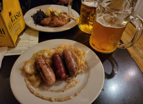 A plate with three types of sausages on sauerkraut, with a side of beer and a meal in the background.