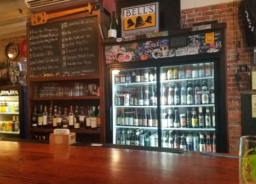 A bar with a wooden counter, shelves of liquor, and a fridge filled with various bottled beers.