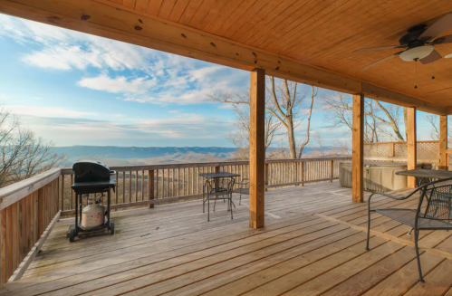 A spacious wooden deck with a grill and tables, overlooking a scenic mountain view under a blue sky.
