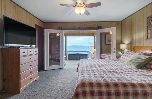 Cozy bedroom with a plaid bedspread, dresser, TV, and doors opening to a deck with a view of the lake.