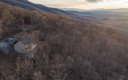 Aerial view of a house on a hillside surrounded by bare trees and distant mountains under a soft sunset sky.