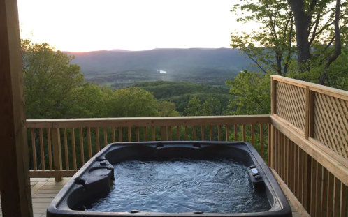 A hot tub on a wooden deck overlooks a scenic valley and mountains at sunset.