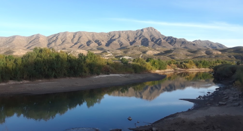 A serene river reflects mountains under a clear blue sky, surrounded by lush greenery.