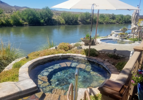 A serene riverside view featuring hot tubs, lush greenery, and a large umbrella for shade.