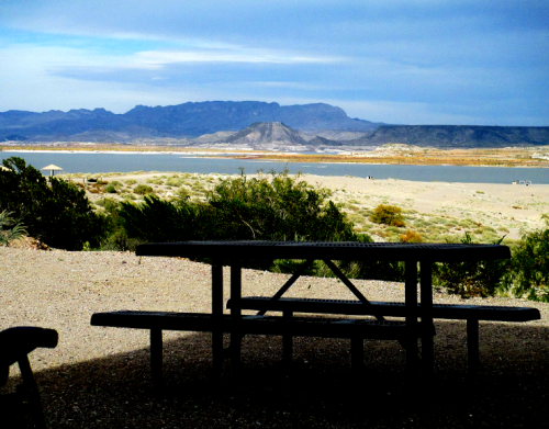 A picnic table in the foreground with a scenic view of mountains and a lake in the background under a cloudy sky.
