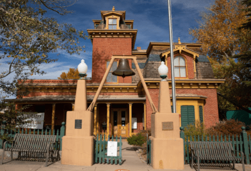 Historic brick building with a tower, bell, and decorative trim, surrounded by benches and autumn foliage.