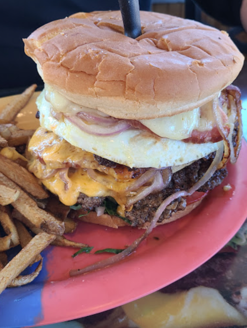 A towering burger with layers of cheese, bacon, and an egg, served with a side of fries on a colorful plate.