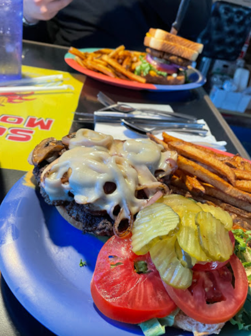 A close-up of a burger topped with cheese, onions, and pickles, served with fries and a side salad on a colorful table.