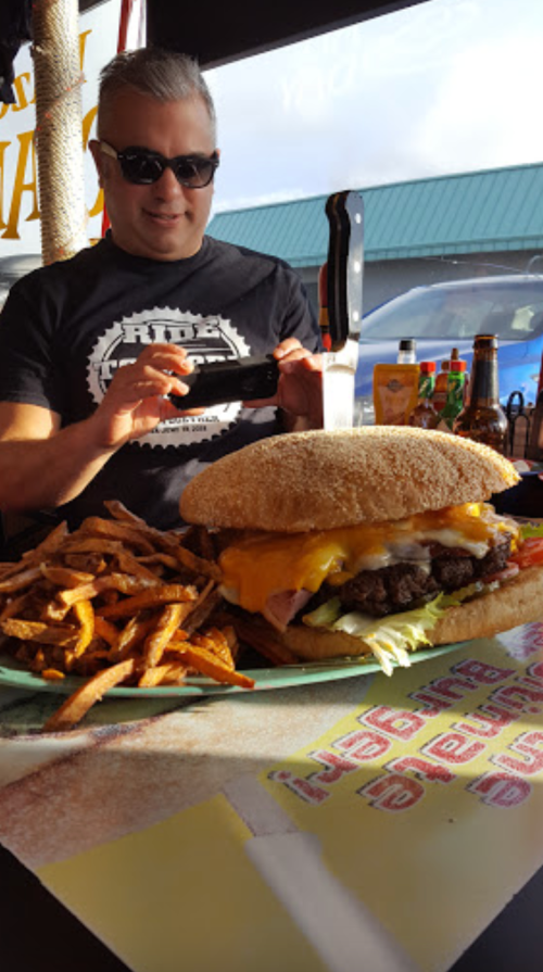 A man takes a photo of a large burger with cheese and fries on a plate at a restaurant.