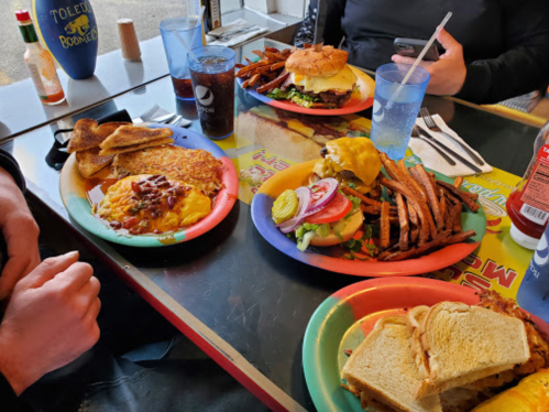 A table with colorful plates of food, including a burger, fries, an omelet, and toast, with drinks in the background.