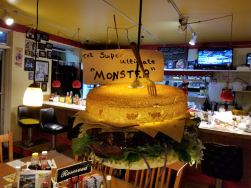 A giant burger display hangs in a diner, labeled "The Super Ultimate Monster," with a fork sticking out of it.