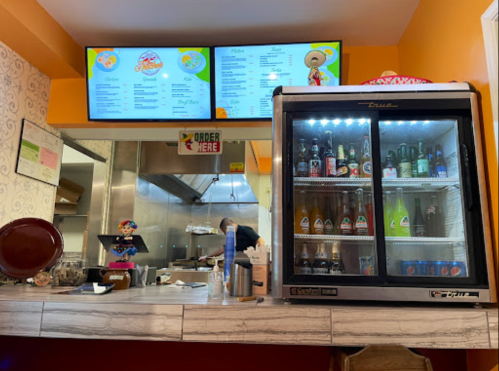 A restaurant counter with a menu on screens, a glass fridge filled with drinks, and a kitchen in the background.