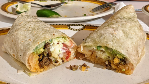 Two burritos cut in half, revealing layers of rice, beans, meat, and fresh toppings on a plate with lime and utensils.