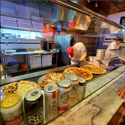A pizza counter with various pizzas and drinks, featuring chefs preparing food in the background.