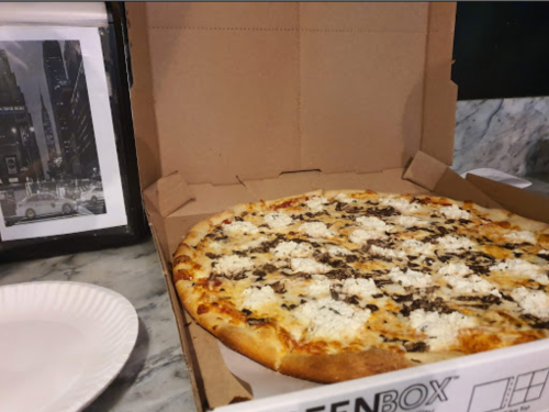 A pizza with white cheese and black toppings in an open pizza box, next to a paper plate and a framed photo.