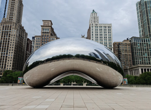 A large, reflective sculpture known as "The Bean" in a city park, surrounded by tall buildings and a cloudy sky.