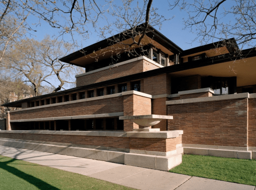 A modernist brick building with large overhangs and a landscaped lawn, surrounded by trees under a clear blue sky.