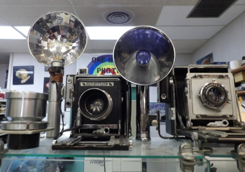 A display of vintage cameras and photography equipment on a glass shelf, featuring various lenses and light sources.