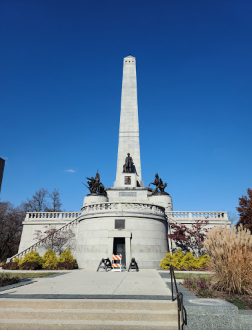 A tall monument with an obelisk, flanked by statues, set against a clear blue sky and surrounded by greenery.